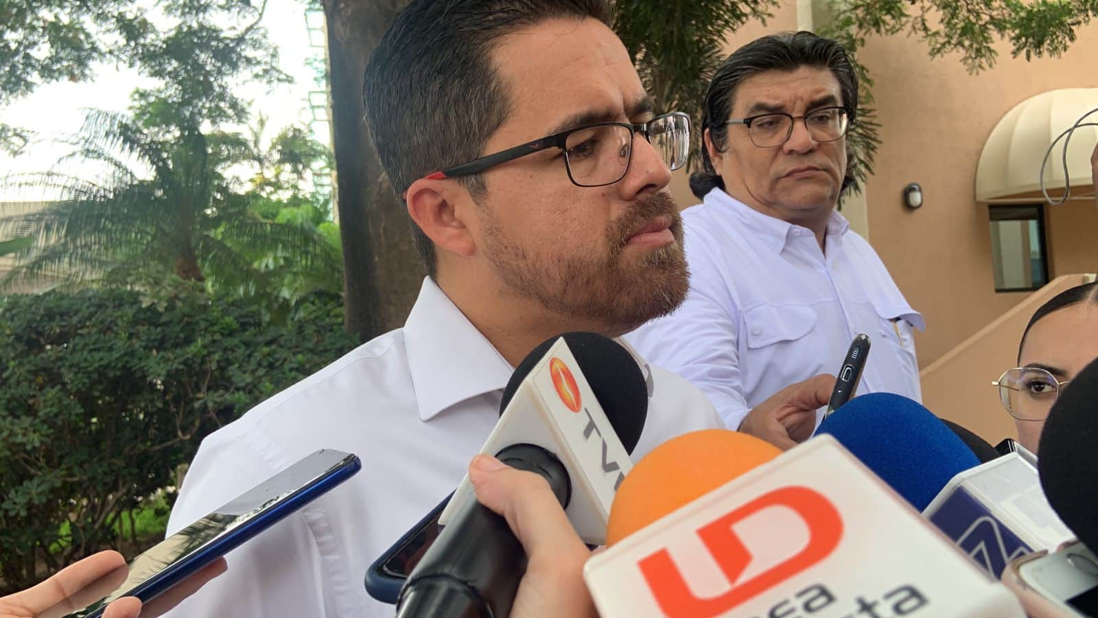 Una persona con lentes hablando con los medios de comunicación, micrófonos