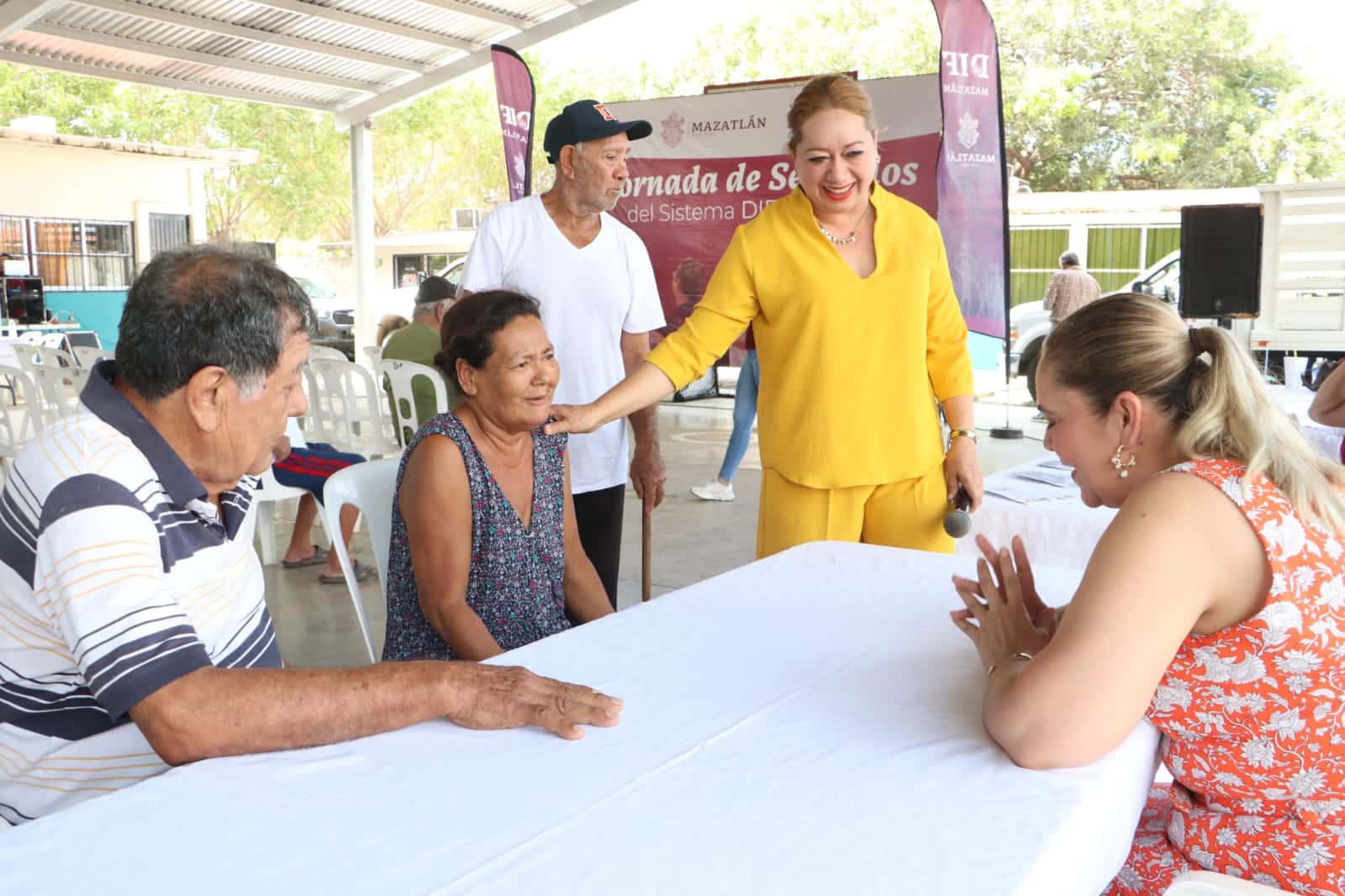 Varias personas sentadas y otras patadas bajo la techumbre de una cancha y una mesa blanca