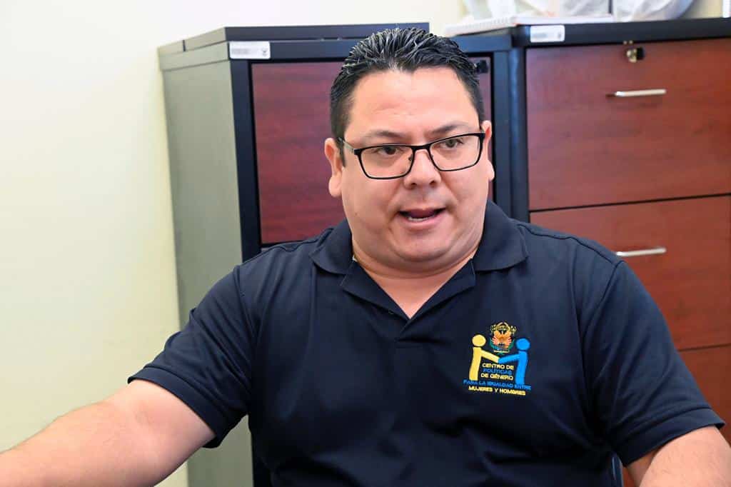Una persona con lentes sentado hablando y unos tipo lockers atrás