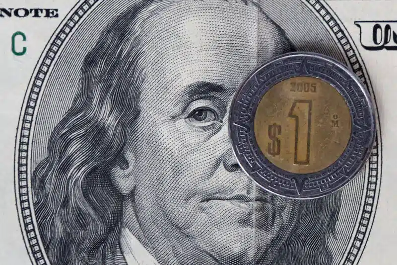 Una moneda de peso mexicana y atrás una parte de un dólar con la imagen de la cabeza de un hombre