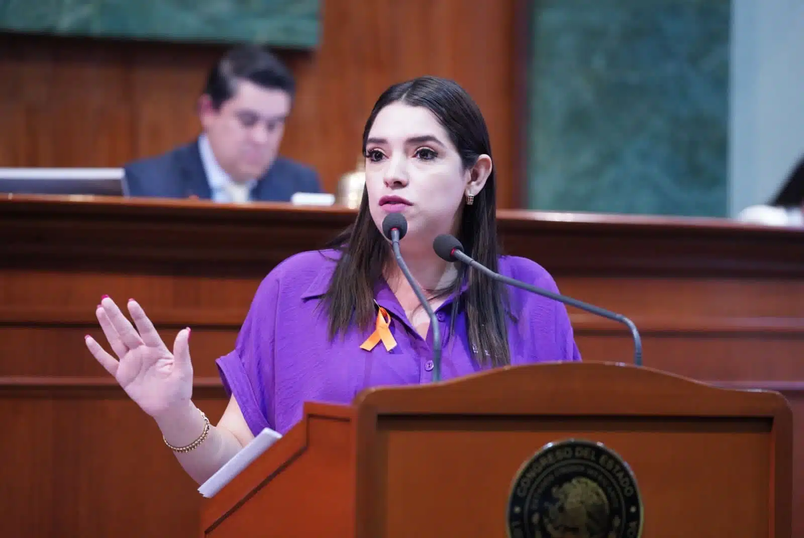 Una persona hablando parada, un micrófono de podium y otra atrás de ella sentada