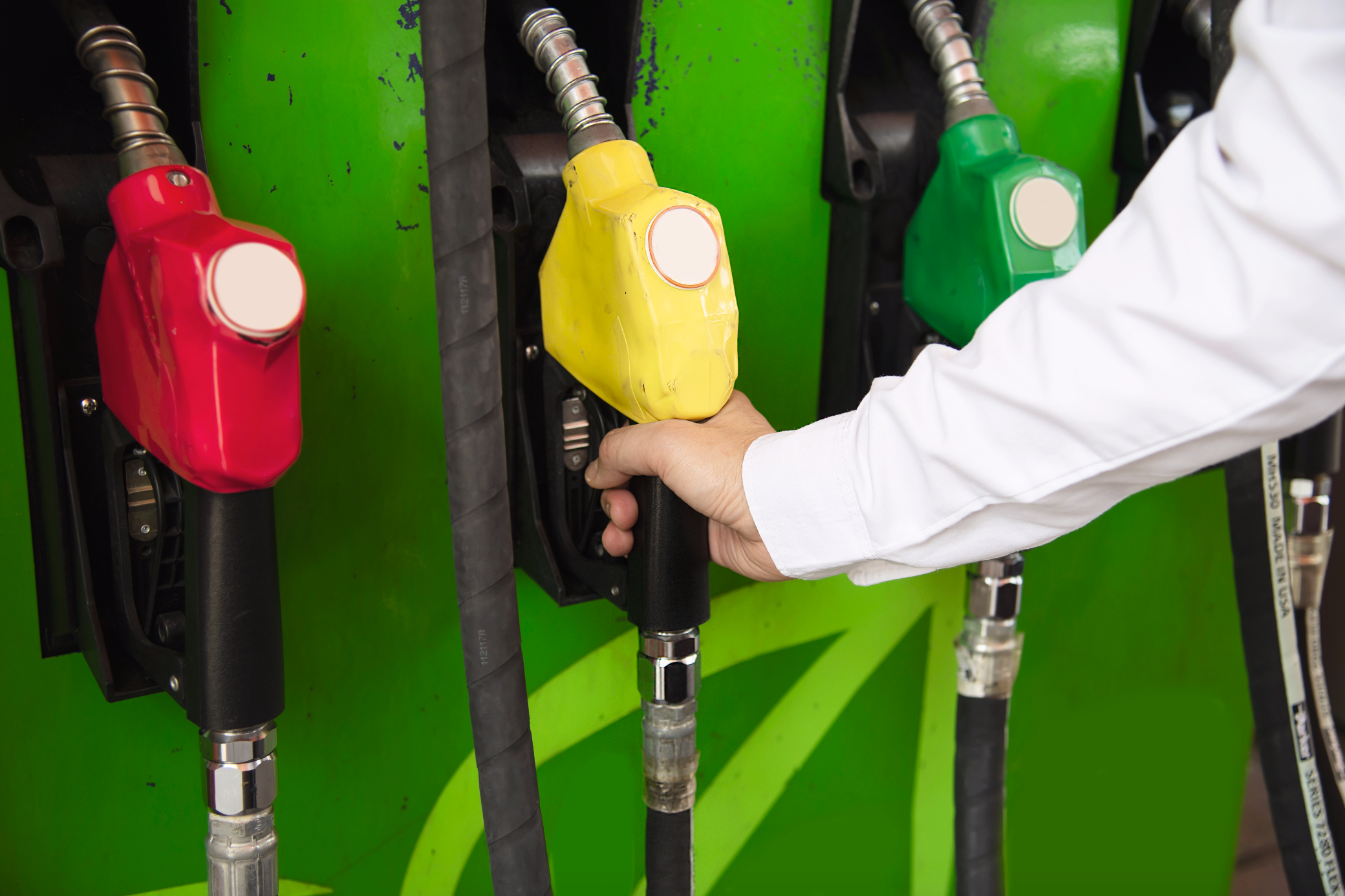 Bombilla de una estación de gasolina y 3 dispensadores de combustibles uno de color verde, otro amarillo y otro rojo, con una mano agarrando una