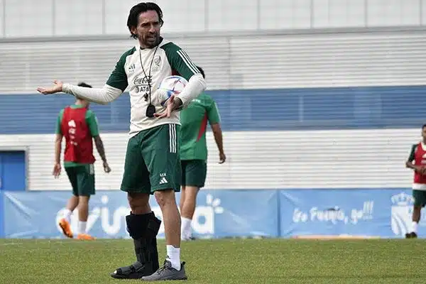 Una persona con las manos extendidas, un balón entre el brazo y el abdomen, con una bota estática en la pierna y un silbato colgando en el pecho, y 3 personas al fondo