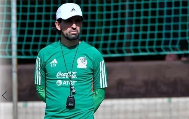 Una persona con las manos hacia atrás, con una gorra y un silbato colgando en el pecho, al fondo una red de portería