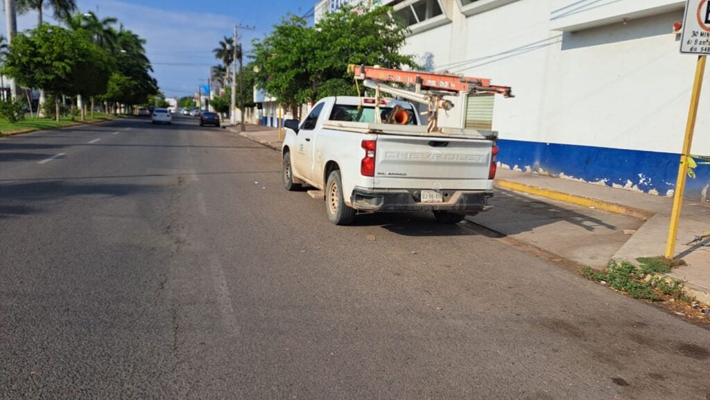 Una camioneta de la CFE estacionada, carros, calle pavimentada, árboles, palmeras y césped