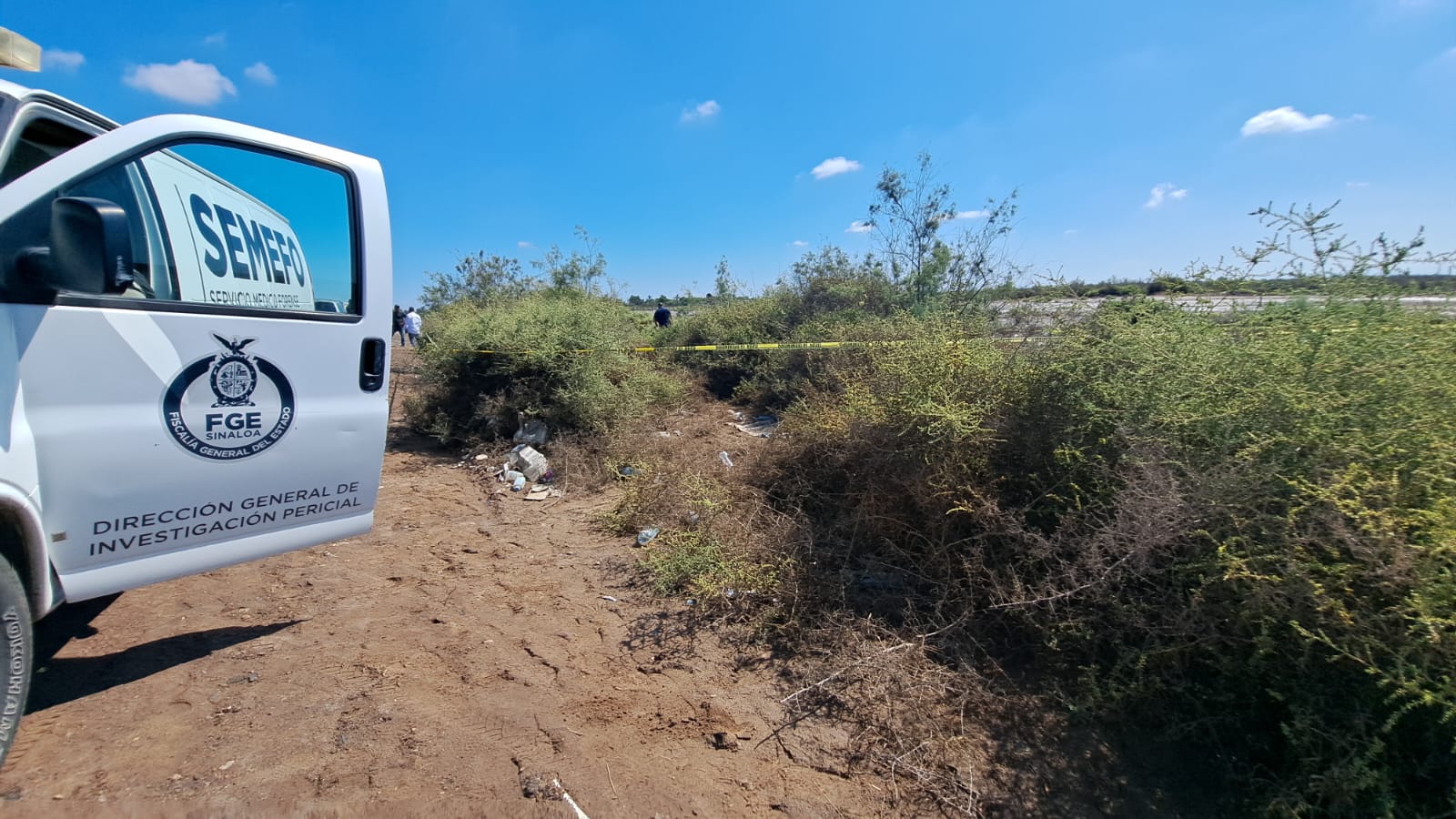 Camioneta con la puerta abierta de la Semefo, arbustos, tierra y cinta amarilla delimitando un área