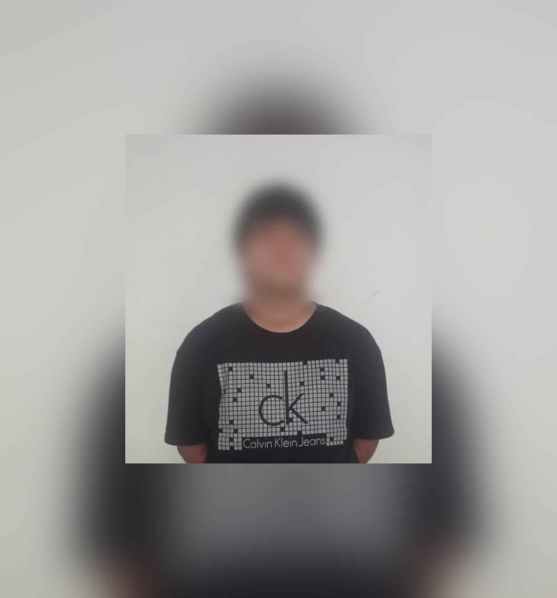 Hombre detenido con playera negra y letras blancas, y la cara tapada