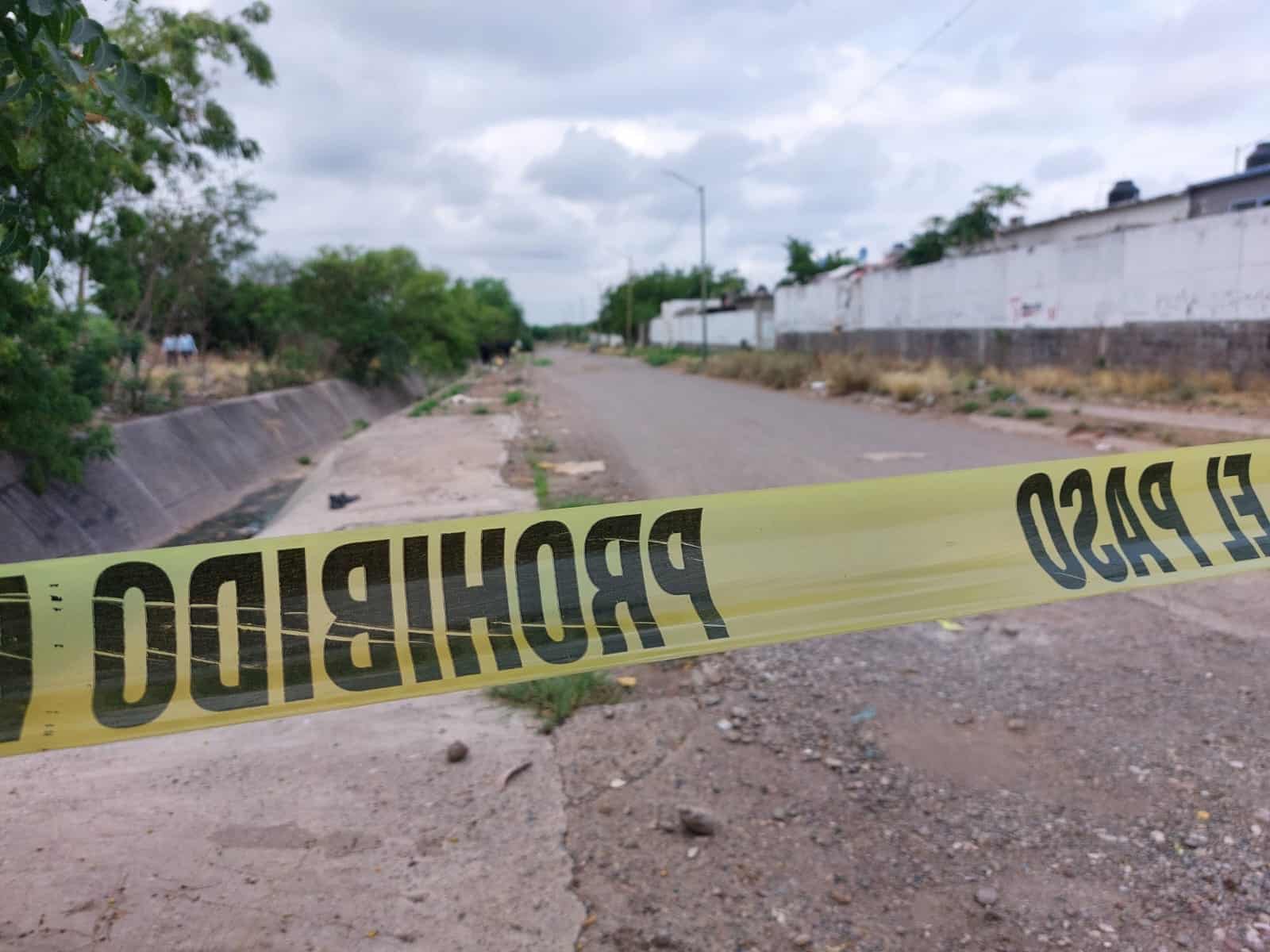 Cinta amarilla delimitando el área de una escena de un crimen, una calle, poste de luz, una barda y árboles junto a un dren