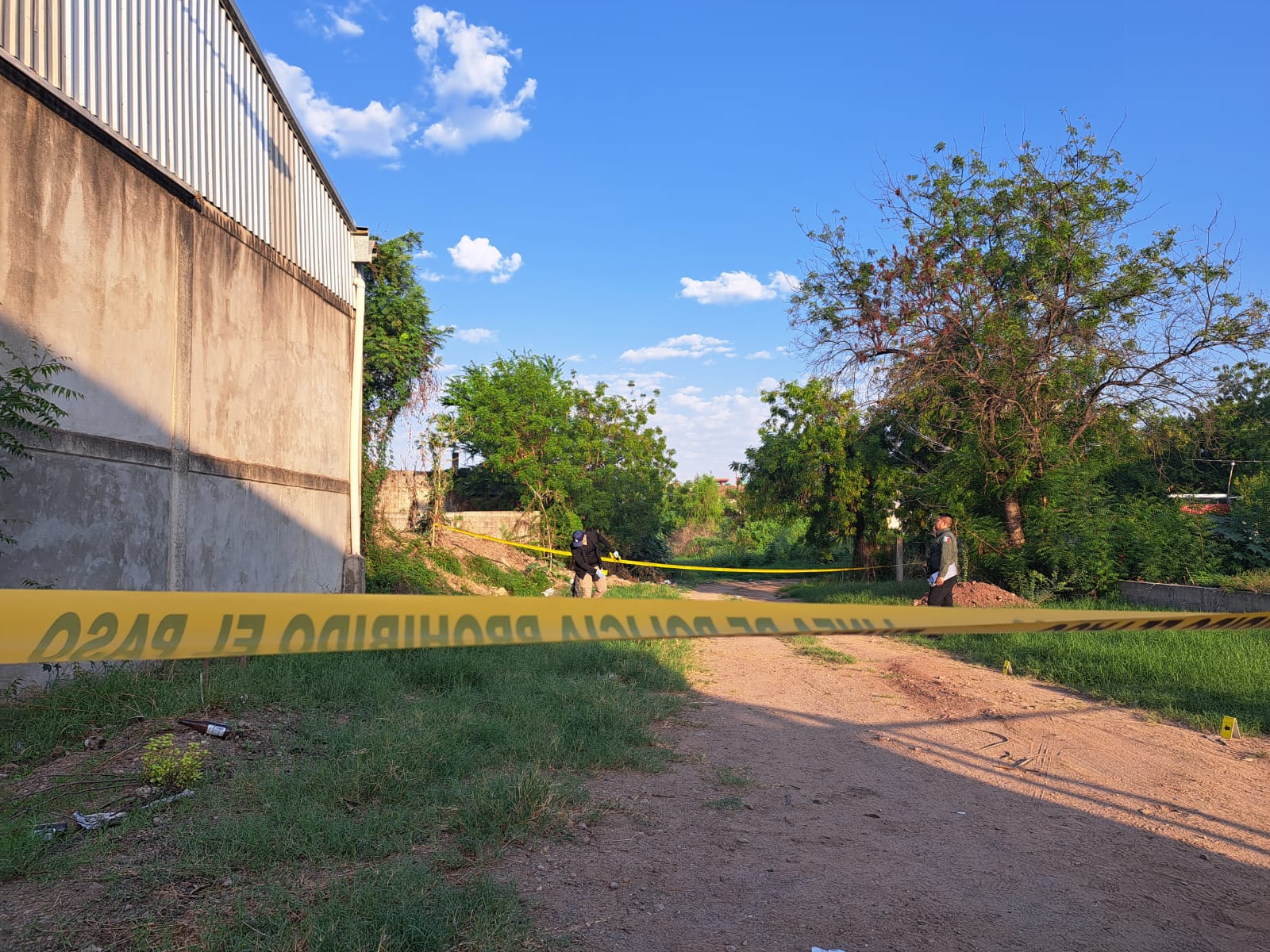 Cinta amarilla delimitando una escena de un crimen, personas en el área, arbustos, árboles, una calle de terracería y una pared de una bodega