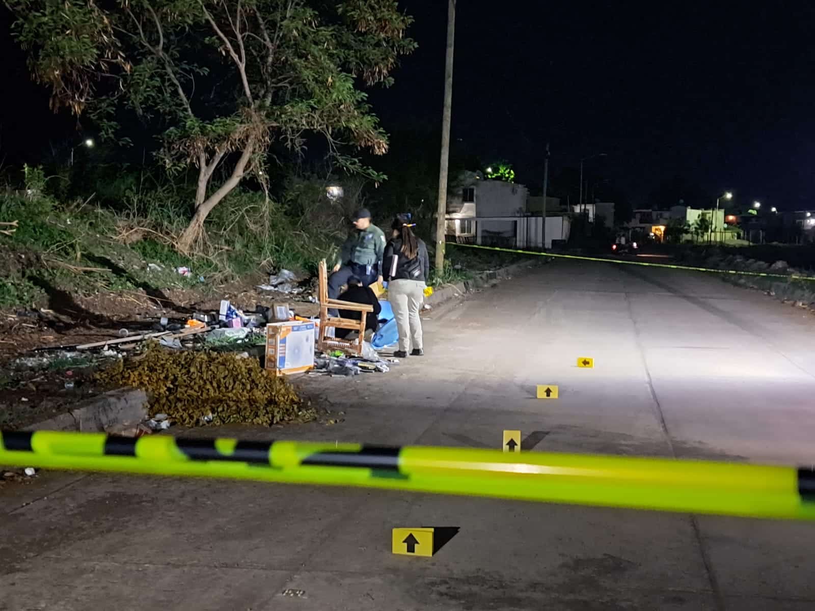 Calle pavimentada, casas al fondo, árboles, ramas, una silla, basura, un tambo azul, cinta amarilla delimitando el área de la escena del crimen, 4 piezas de señalización serigrafiadas con flechas de color amarillo y 3 personas cerca del tambo
