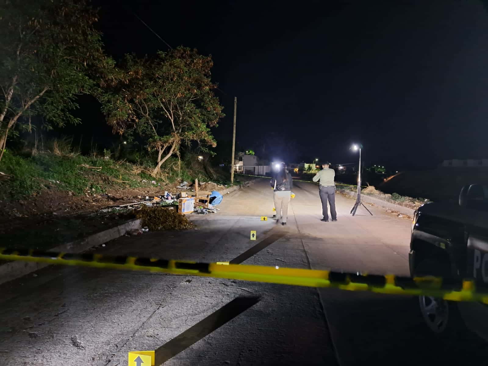 Calle pavimentada, casas al fondo, árboles, ramas, un tripié de luz, una silla, basura, un tambo azul, cinta amarilla delimitando el área de la escena del crimen, 2 piezas de señalización serigrafiadas con flechas de color amarillo, la parte delantera de una camioneta y 2 personas cerca de la escena