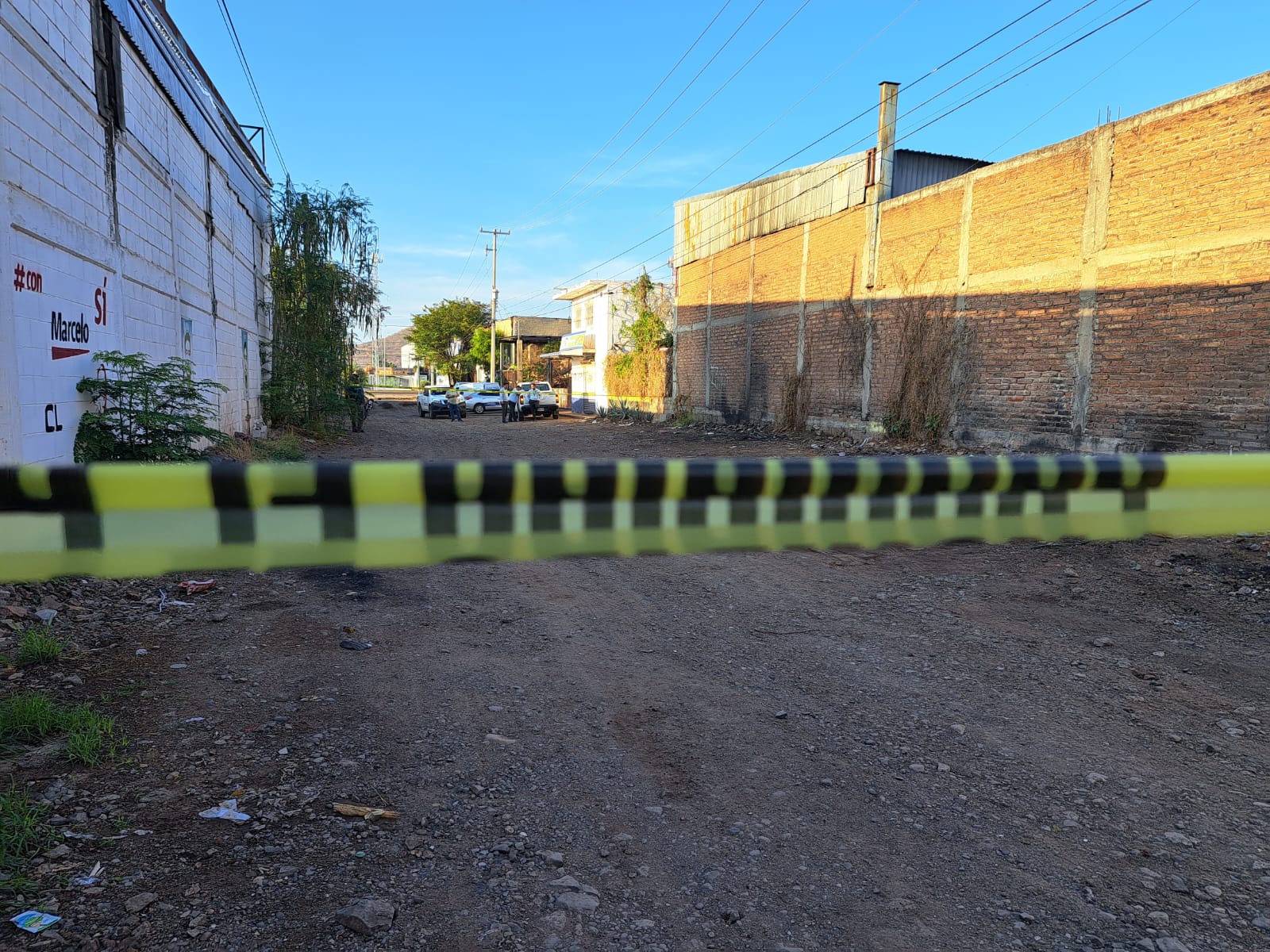 Calle de terracería, área delimitada con cinta amarilla, paredes de construcciones, al fondo carros y personas