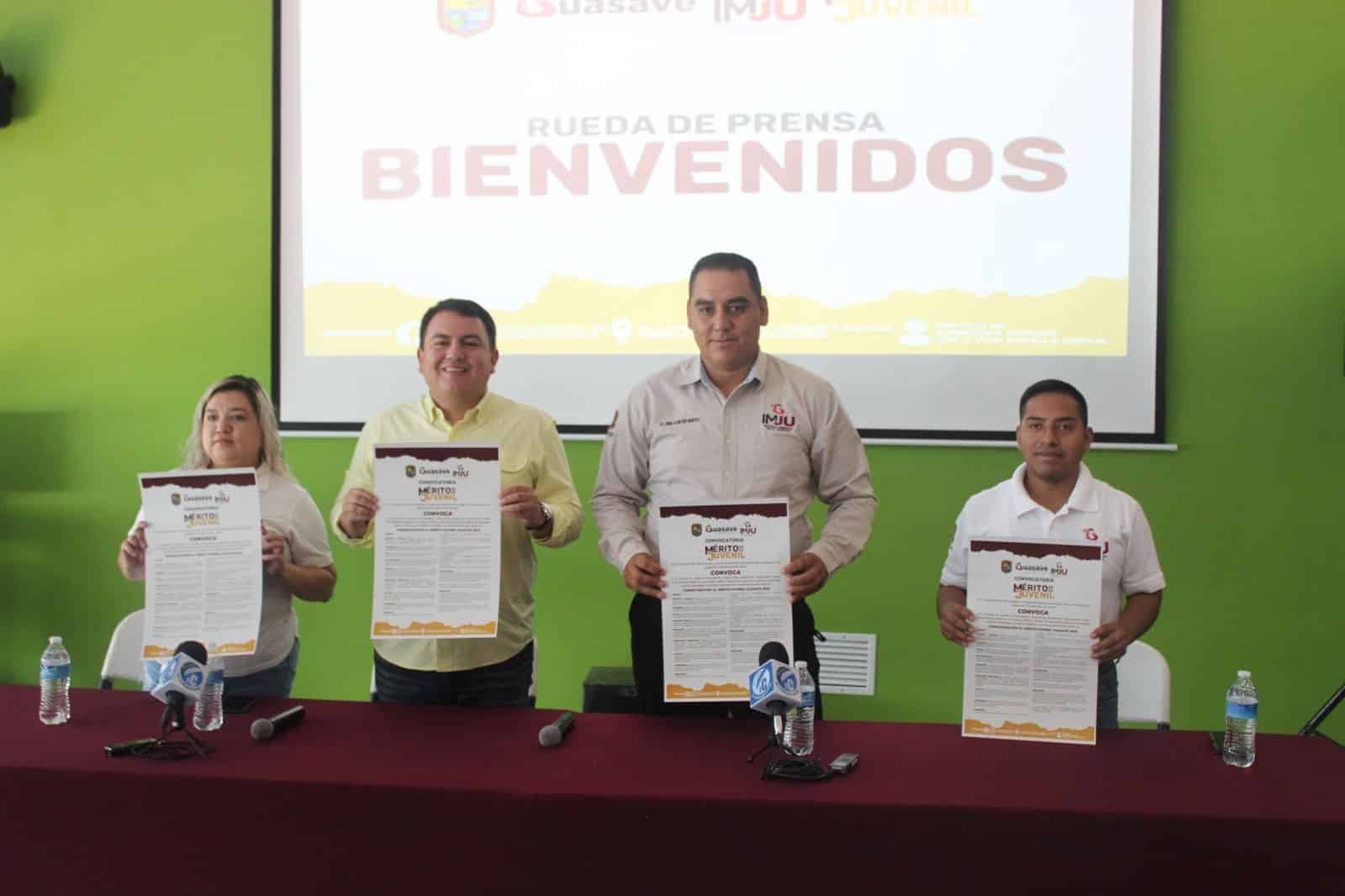 4 personas con un papel en la mano cada uno, una mesa con mantel de color tinto, al fondo una pantalla en donde se proyecta, micrófonos y botellas de agua