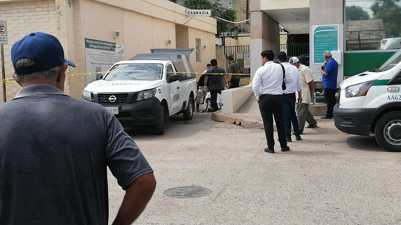 Una camioneta de la Semefo, personal de dicha institución, 5 personas más observando la escena, cinta amarilla delimitando el área, paredes del IMSS y la parte frontal de una ambulancia del instituto