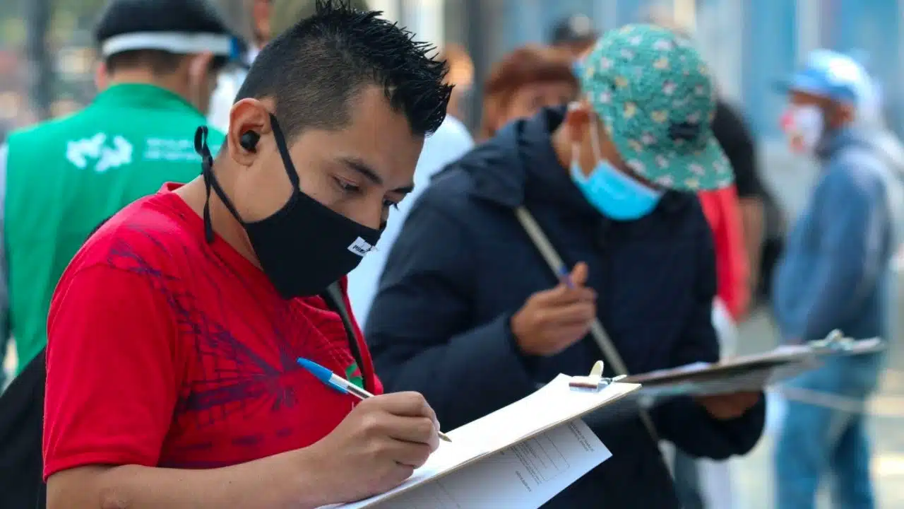 2 personas con cubrebocas y una de ellas con gorra, 2 plumas, 2 tablas en las que escriben, hojas de papel y al fondo varias personas