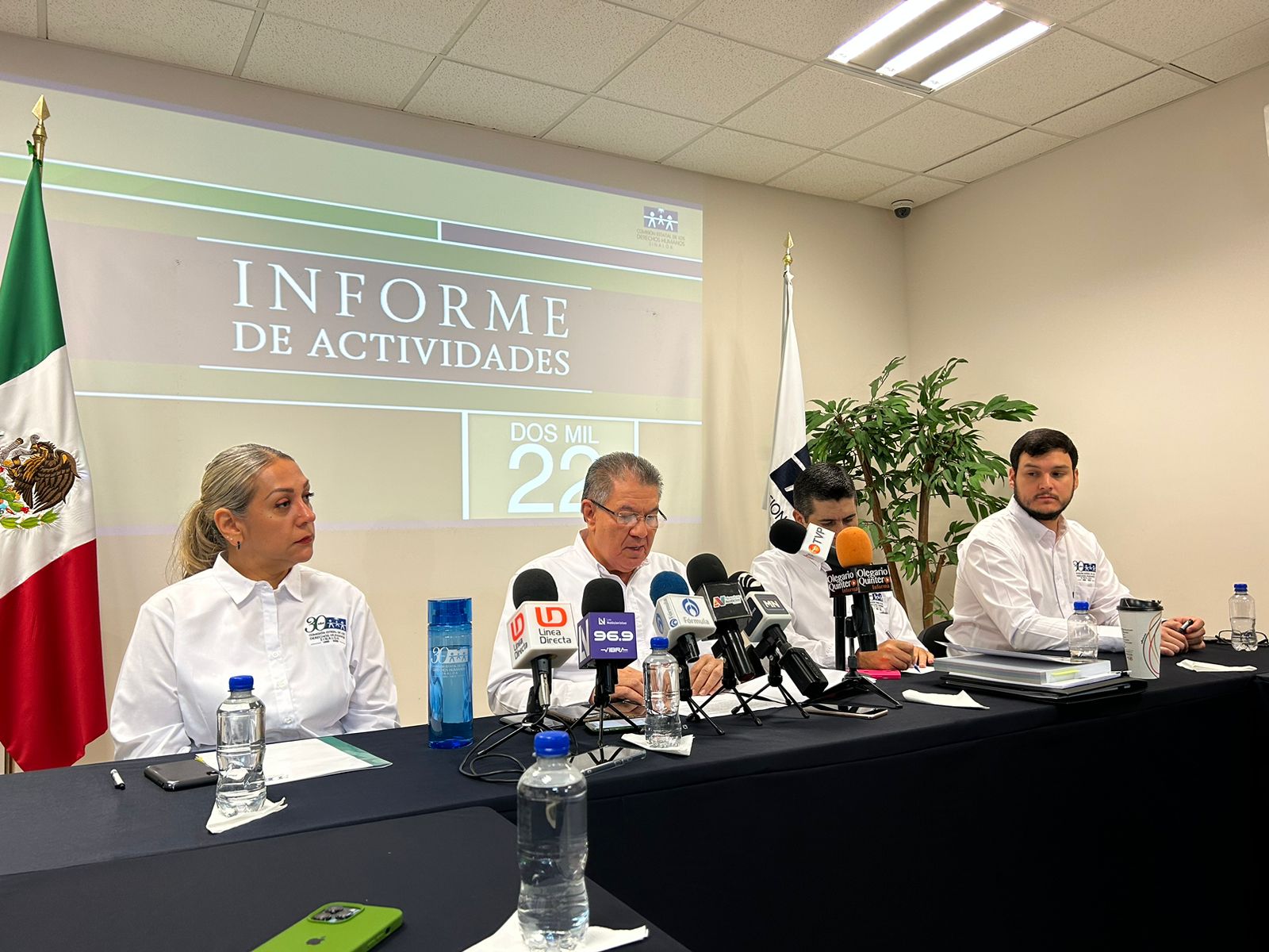 4 personas sentadas, una mesa larga, 7 micrófonos de medios de comunicación, 2 banderas una de México y otra de color blanco, botellas de agua y una pared que está proyectado unas letras