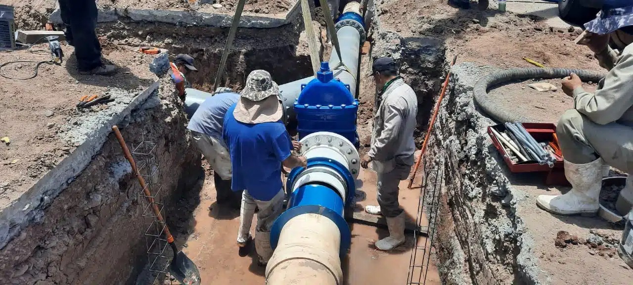 Un tubo grande subterráneo, 3 personas arreglándolo, una más en cuclillas mirando desde arriba, los pies de otra, varillas, agua, lodo, tubos, tierra, pala y escombros