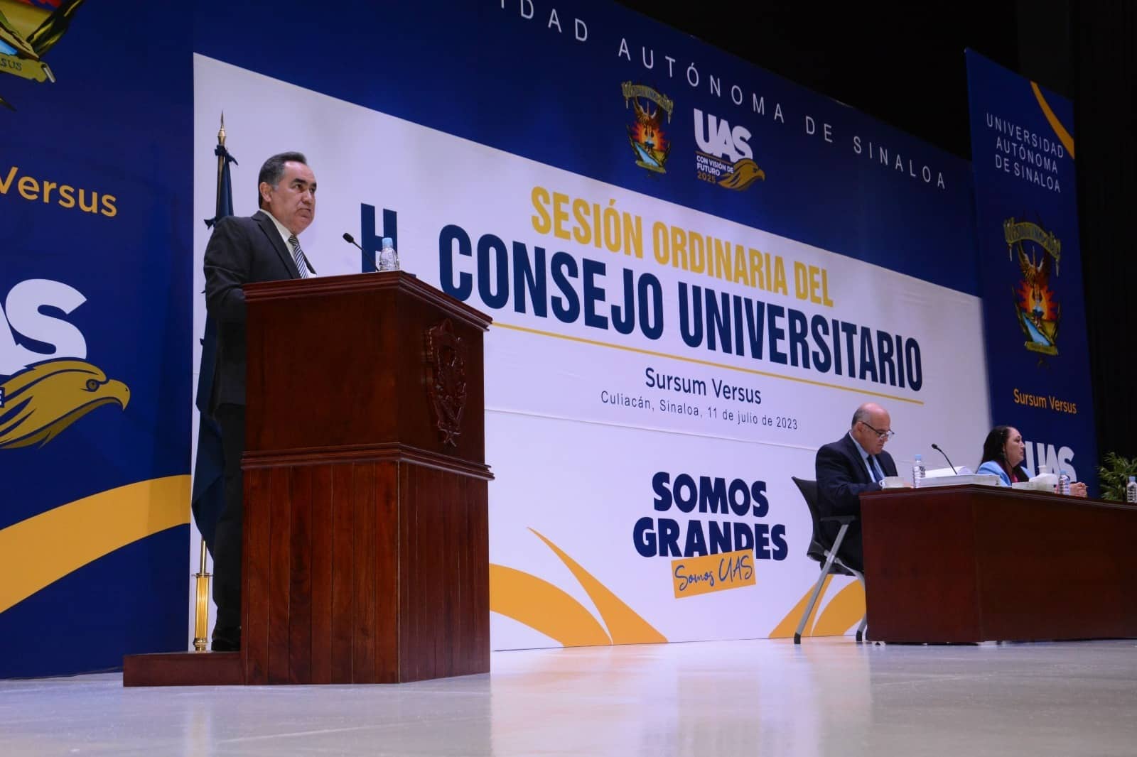 Una persona en un podium, 2 más sentadas, una mesa y al fondo una lona