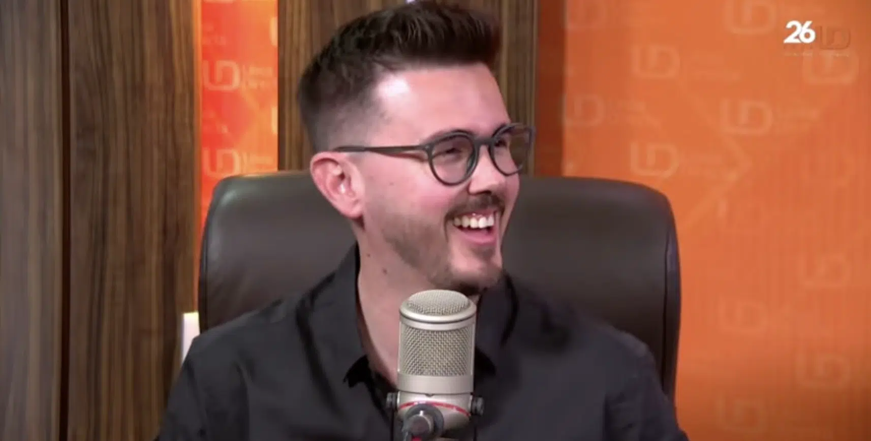 Una persona con lentes riéndose sentada y un micrófono de estudio al frente