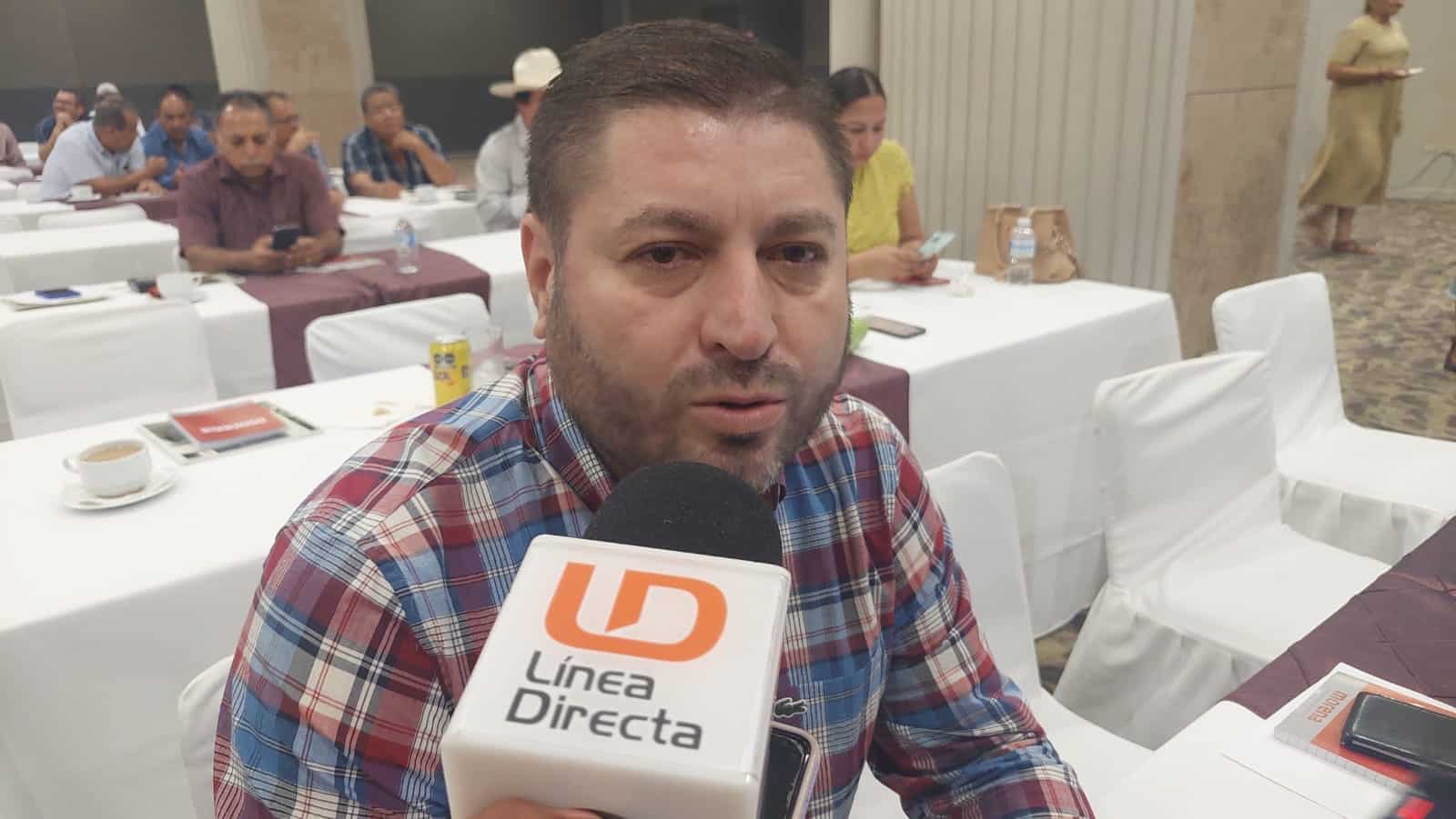 Una persona sentada hablando frente a un micrófono de LD, sillas, mesas y personas al fondo