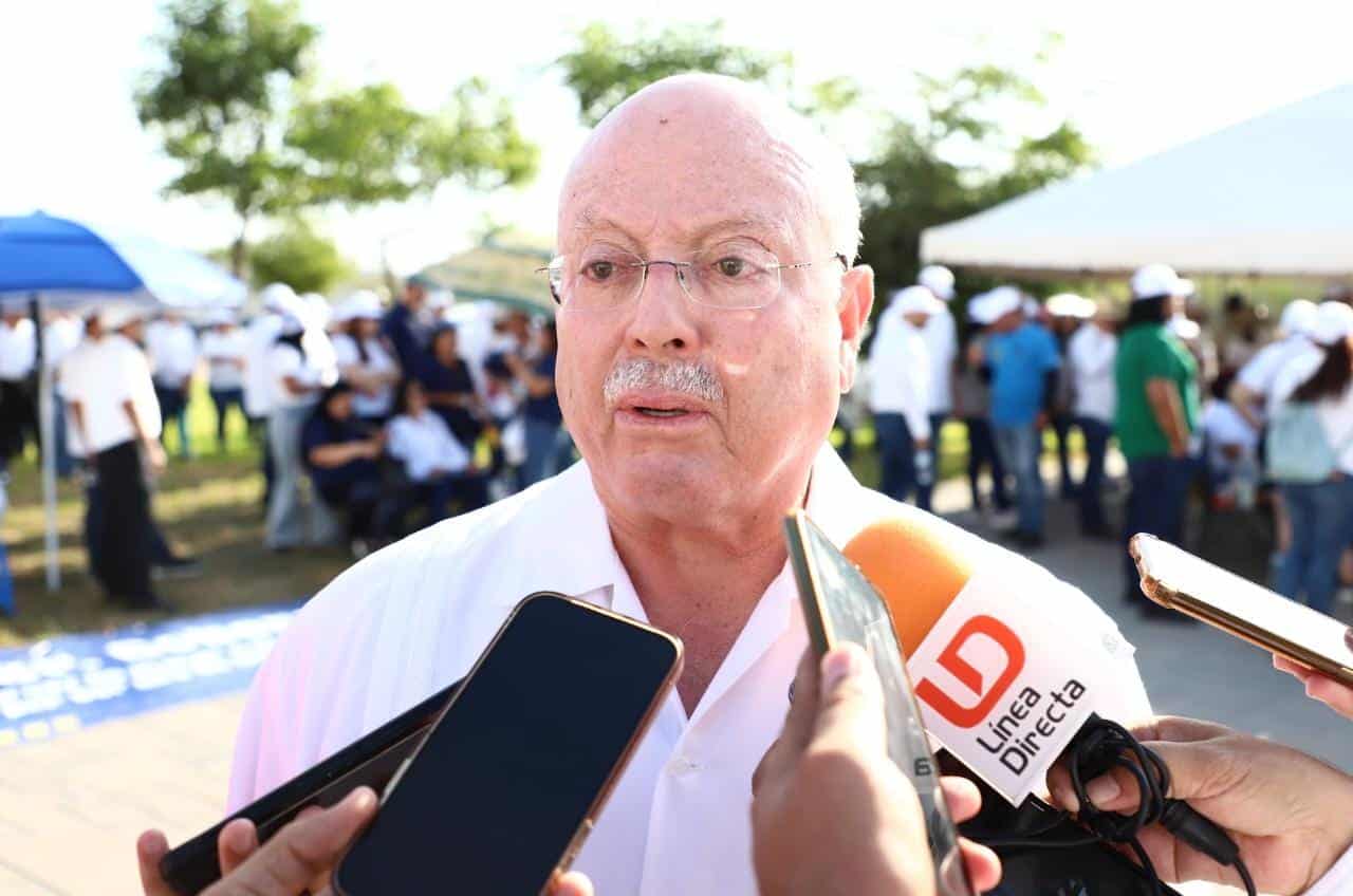 Una persona con lentes hablando ante medios de comunicación, micrófonos y al fondo personas