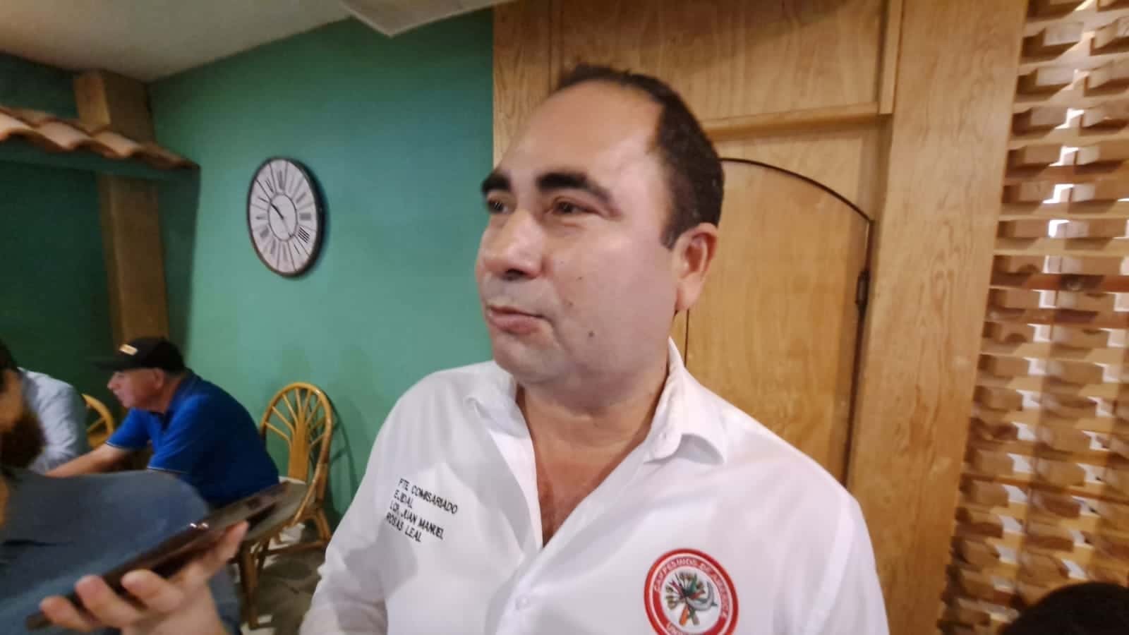 Una persona hablando, una puerta de madera, una silla, un reloj de pared, una persona sentada y un celular