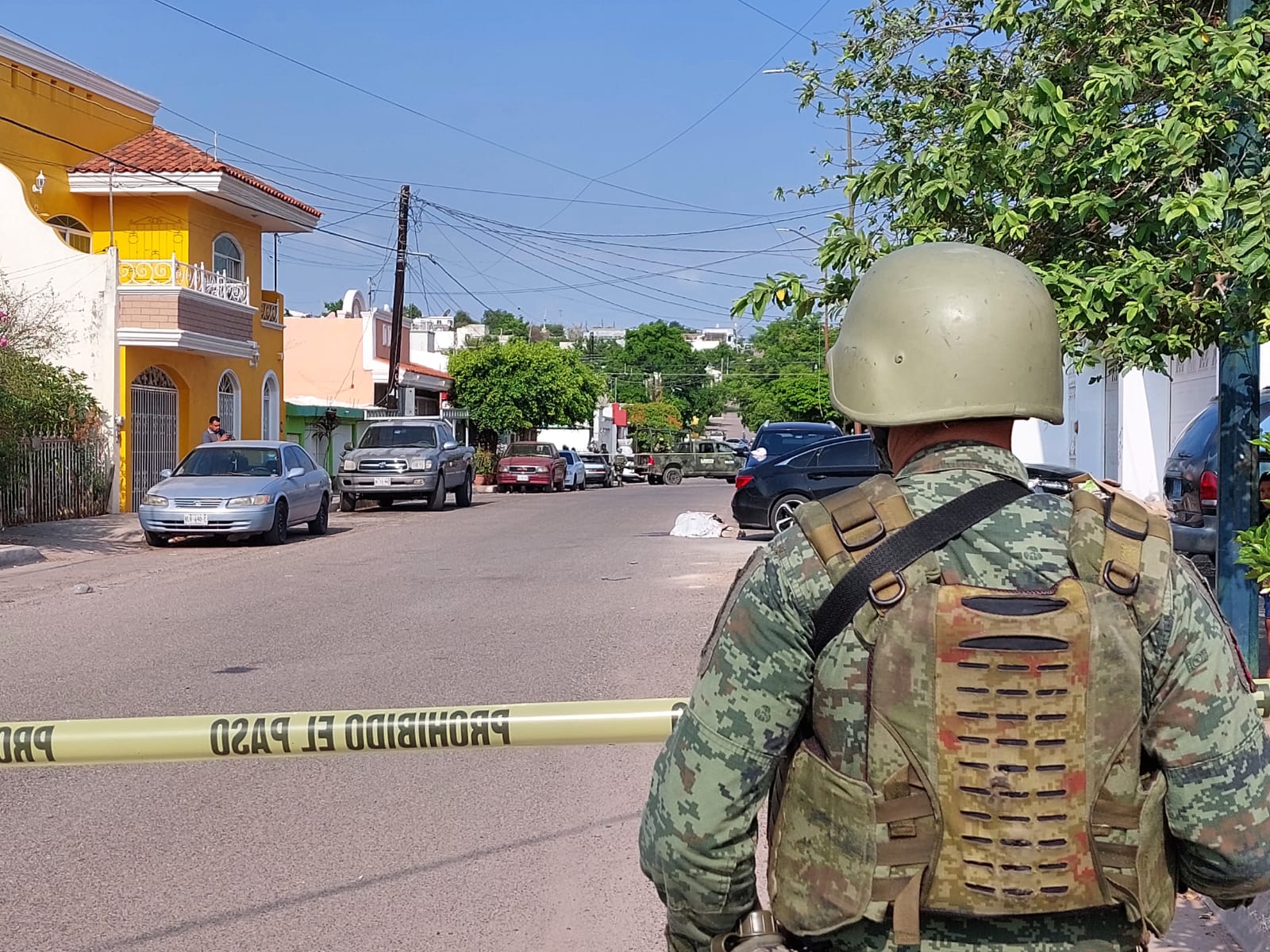 Una persona con uniforme de militar y un casco de espaldas, cinta amarilla delimitando el área, carros estacionados, árboles y casas