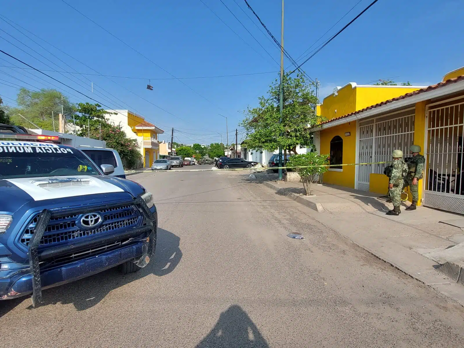 Parte delantera de una camioneta de policía, una calle pavimentada, carros estacionados, unos tenis colgados en unos cables, 2 personas paradas a un lado de una casa, árboles, cinta amarilla delimitando el área y casas