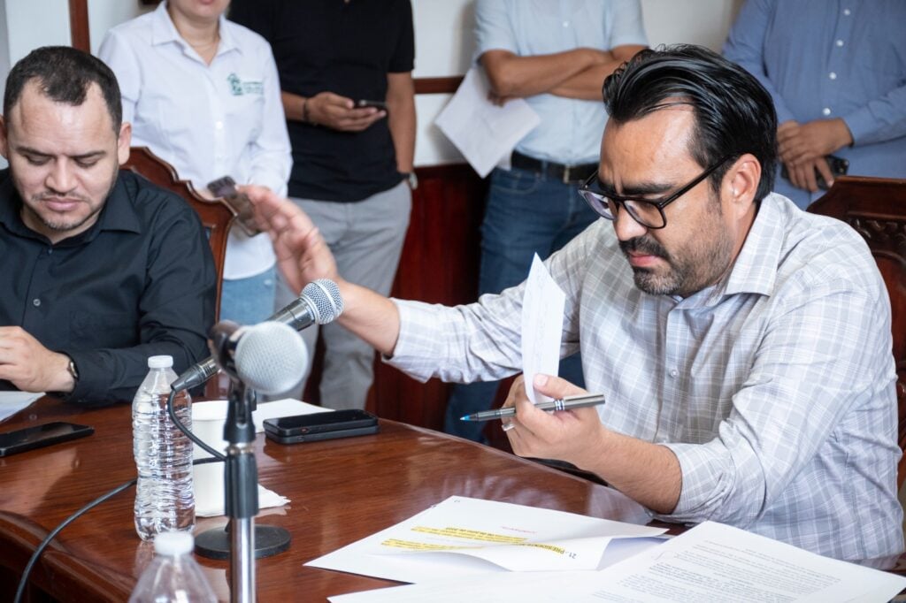 2 personas una de ellas con la mano alzada, en la otra mano una pluma y un papel, papeles sobre la mesa, botella de agua y un micrófono