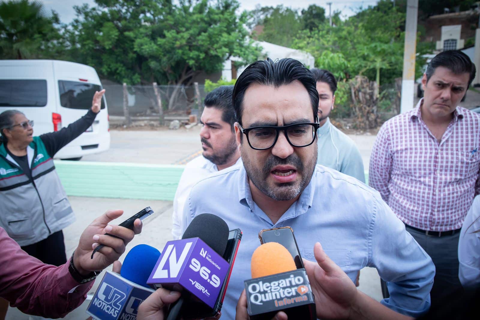 Una persona con lentes hablando ante los micrófonos de los medios de comunicación y personas al fondo