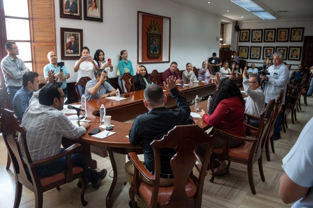 Muchas personas sentadas con la mano alzada, otras paradas, papeles sobre la mesa, botellas de agua y un micrófono, otra persona con una videocámara y al fondo cuadros