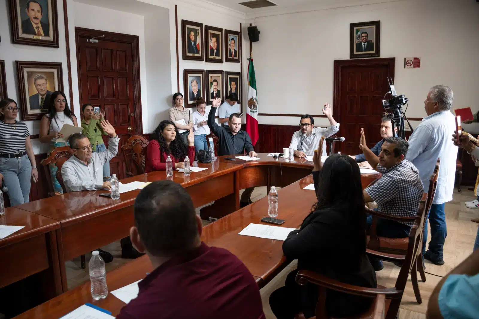 Muchas personas sentadas con la mano alzada, otras paradas, papeles sobre la mesa, botellas de agua y un micrófono, otra persona con una videocámara, al fondo cuadros y una bandera de México