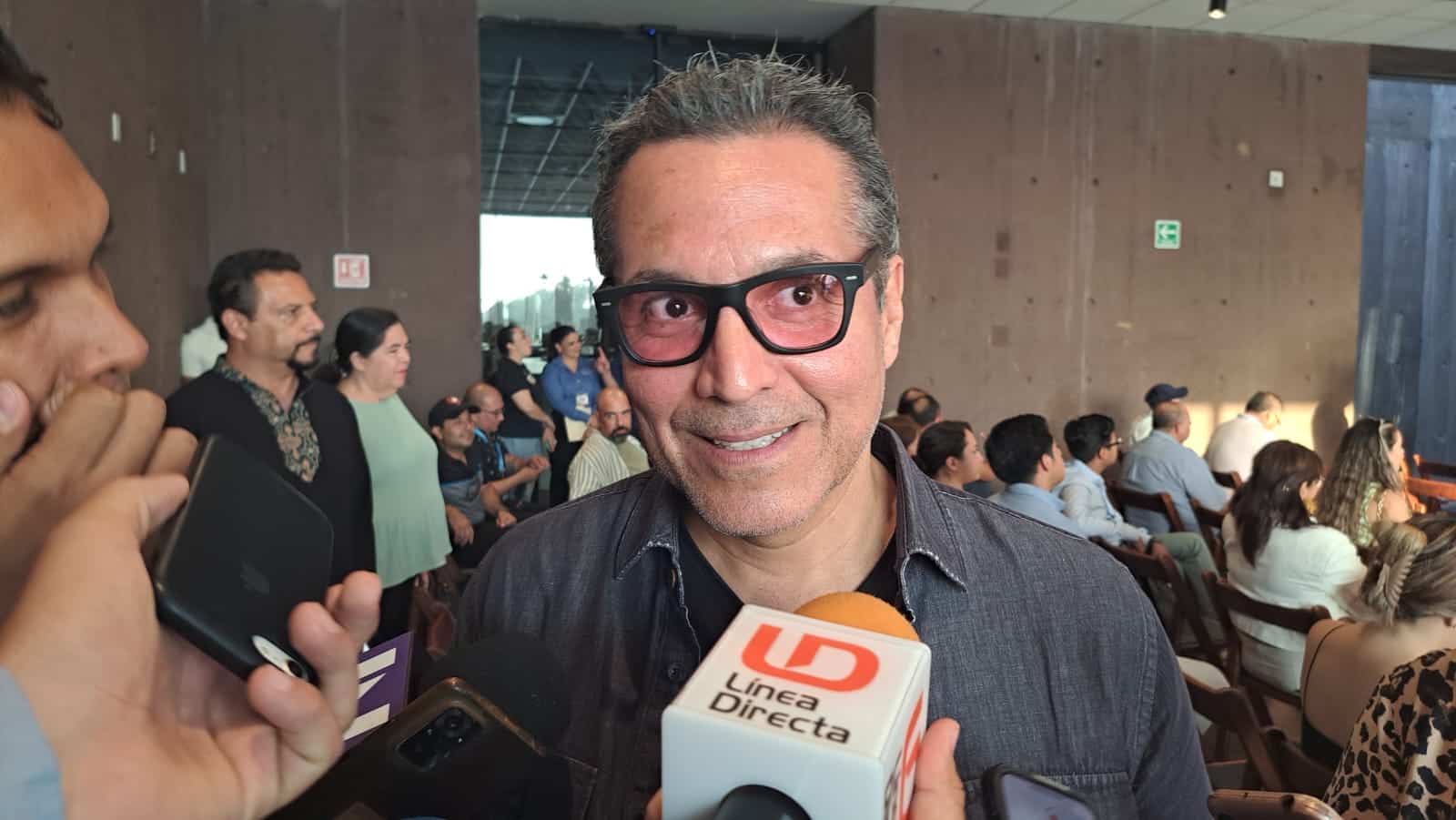 Una persona con lentes, un micrófono de LD, y muchas personas al fondo
