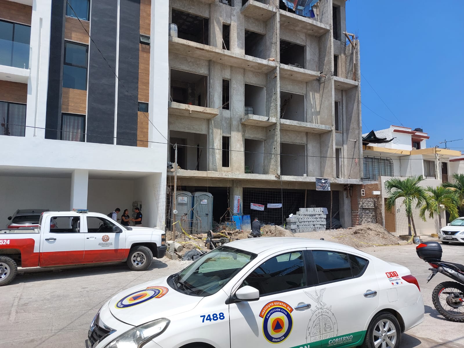 Edificio de departamentos, un carro y una camioneta estacionados, palmeras, casas, escombros y personas