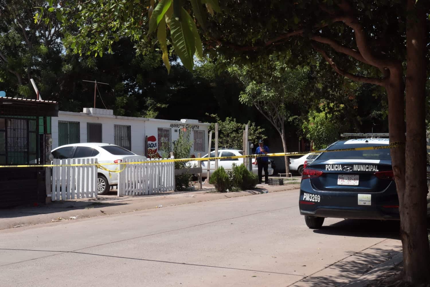 Un carro de la policía sobre una calle pavimentada, casas y carros estacionados, árboles, personas y cinta amarilla delimitando el área de un crimen