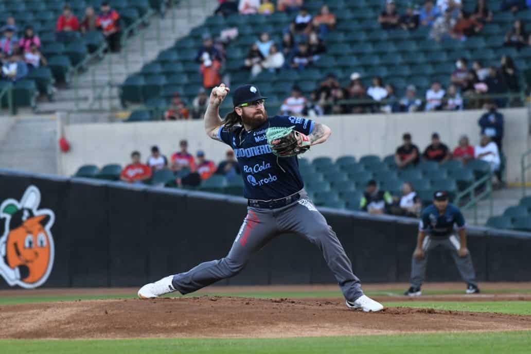 Una persona con gorra, lentes, manilla y una pelota en la mano parada en la lomita de una cancha de beisbol, butacas y personas al fondo
