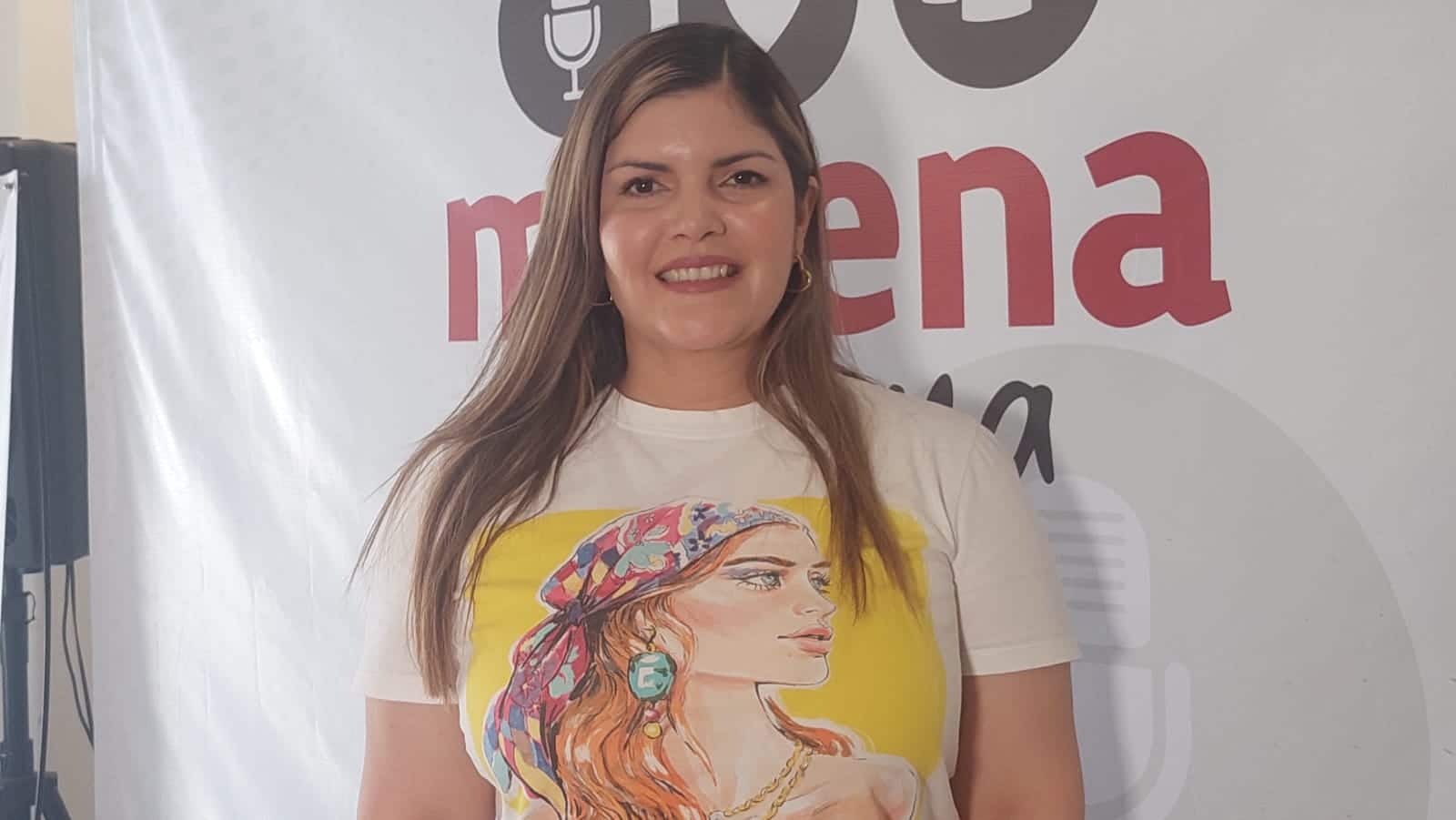 Una mujer sonriendo para una foto, y atrás de ella una lona con letras de un partido político