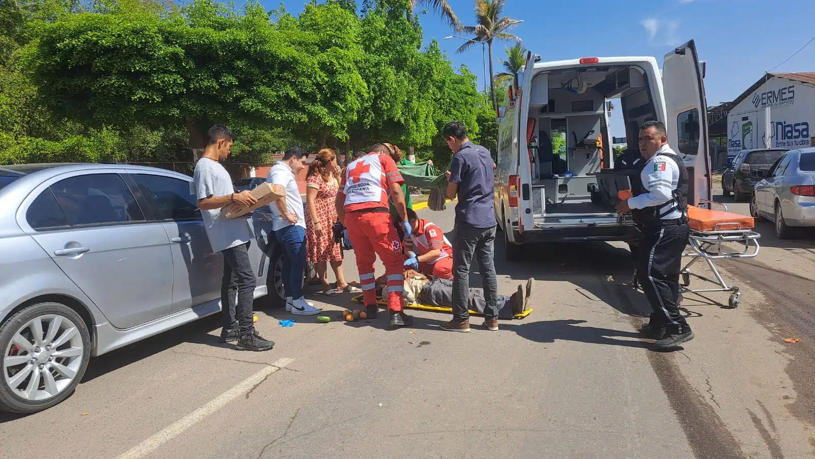 Un carro, una persona en el suelo siendo atendida por 2 más, y 6 más mirando, una camilla, una ambulancia con las puertas abiertas