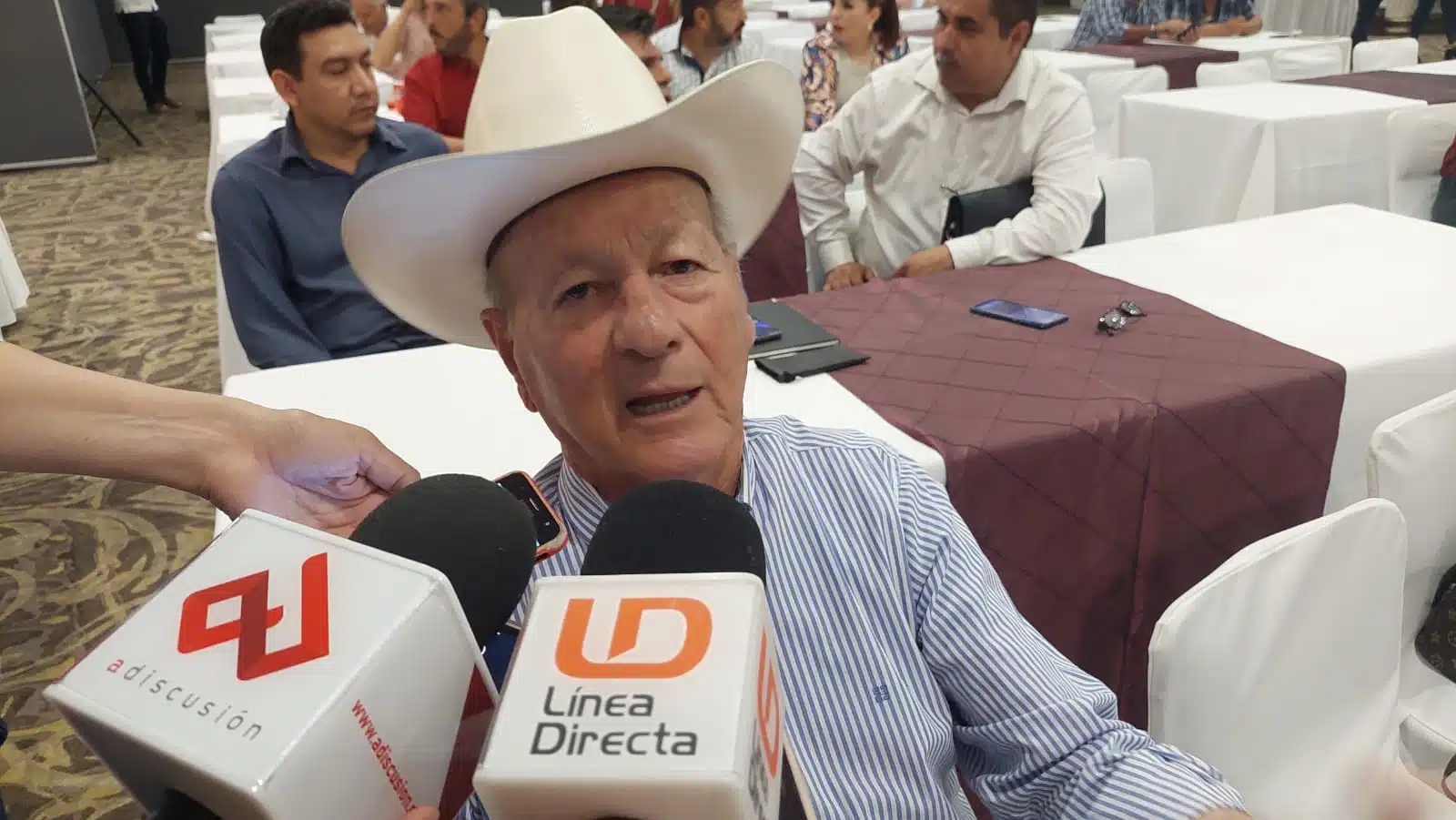 Una persona con sombrero sentada hablando ante 2 micrófonos de medios de comunicación, atrás una mesa con mantel blanco y café, un celular, carpeta y 2 personas