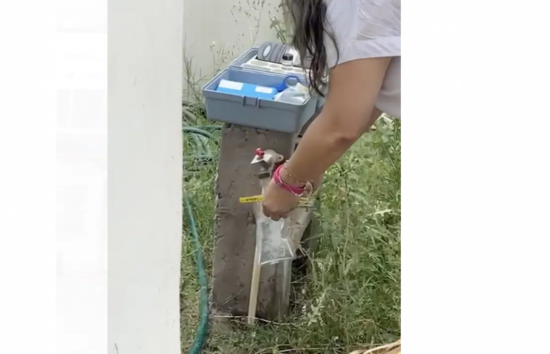 Llave del agua abierta, manos de una persona con una bolsita llenándola del líquido, pasto y una estructura de concreto