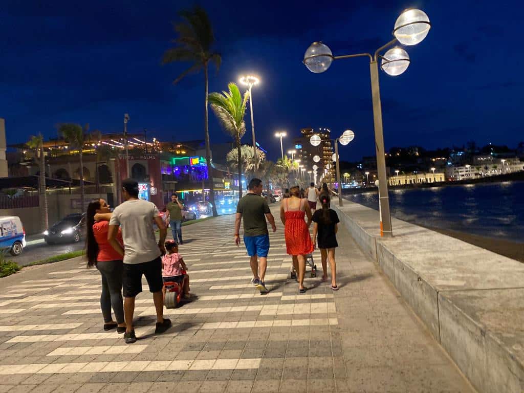 Personas caminando sobre la banqueta, lámparas prendidas, cielo obscuro, agua del mar, luces de carros, edificios y casas