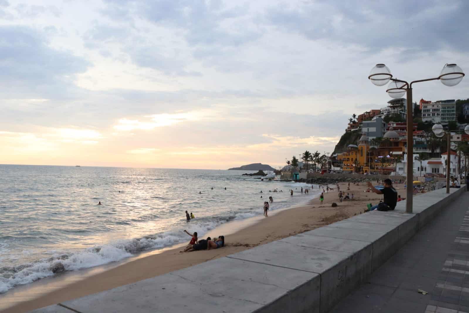 Agua del mar, arena, parte de la banqueta, cielo nublado con un poco de rayos de sol, lámparas, personas y al fondo casas, edificios y palmeras