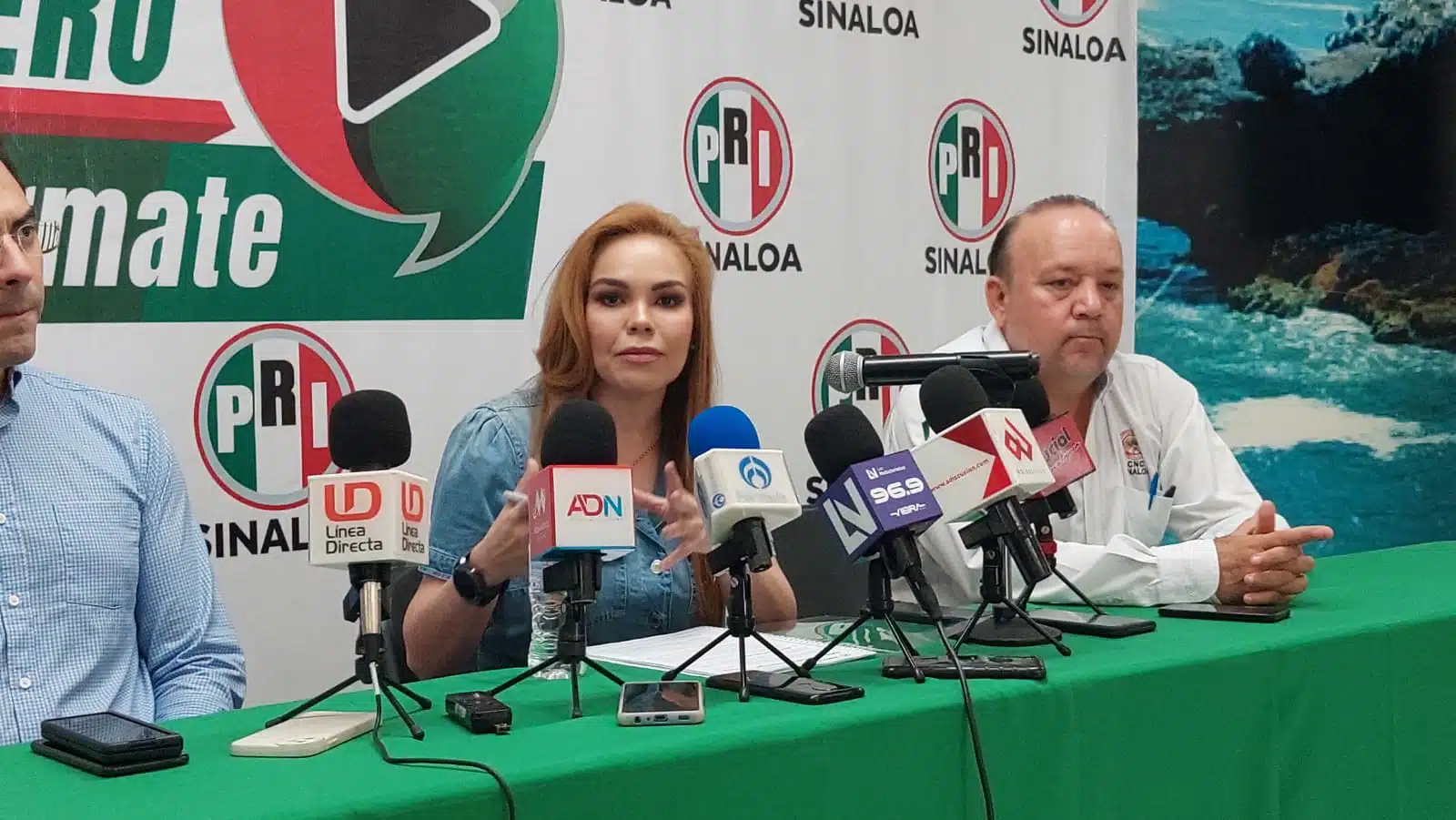 3 personas en una conferencia de prensa, micrófonos, una mea con un mantel de color verde y al fondo una lona con las siglas y colores del PRI