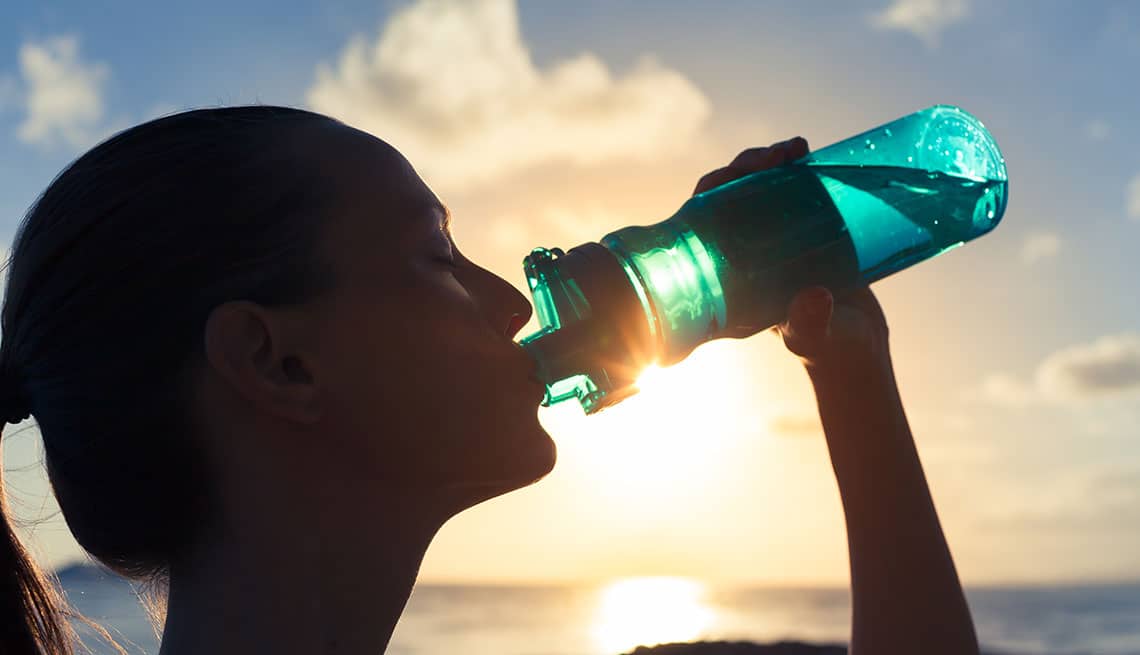 La cabeza de una mujer tomando agua de un bule color azul y al fondo rayos del sol y nubes