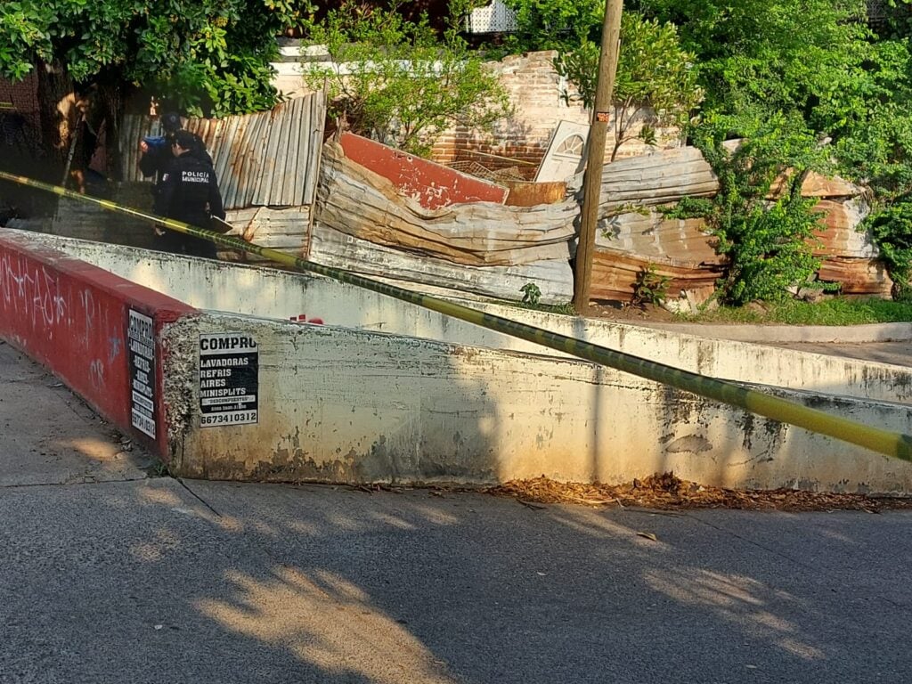 Una pequeña barda de un canal pluvial, 2 personas paradas, cinta amarilla delimitando el área, un poste y láminas