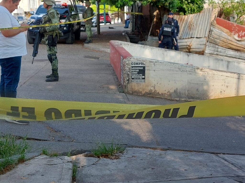 Una pequeña barda de un canal pluvial, 5 personas, cinta amarilla delimitando el área y carros estacionados