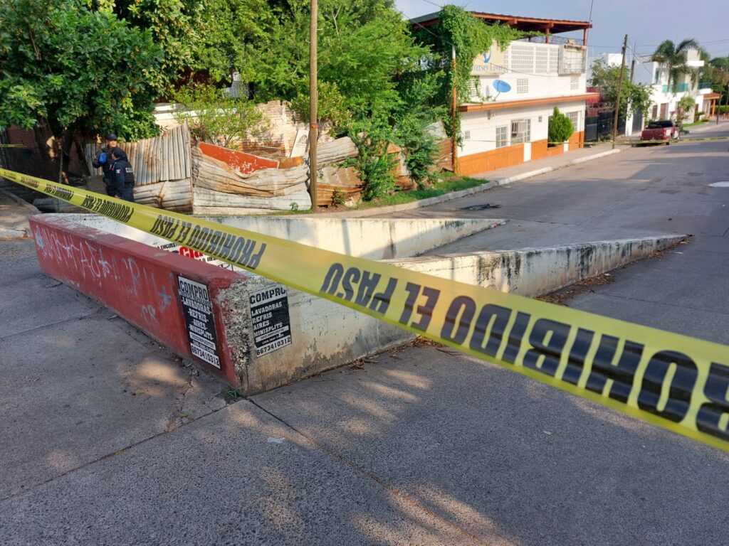 Una pequeña barda de un canal pluvial, 2 personas paradas, cinta amarilla delimitando el área, un poste, árboles, casas y láminas