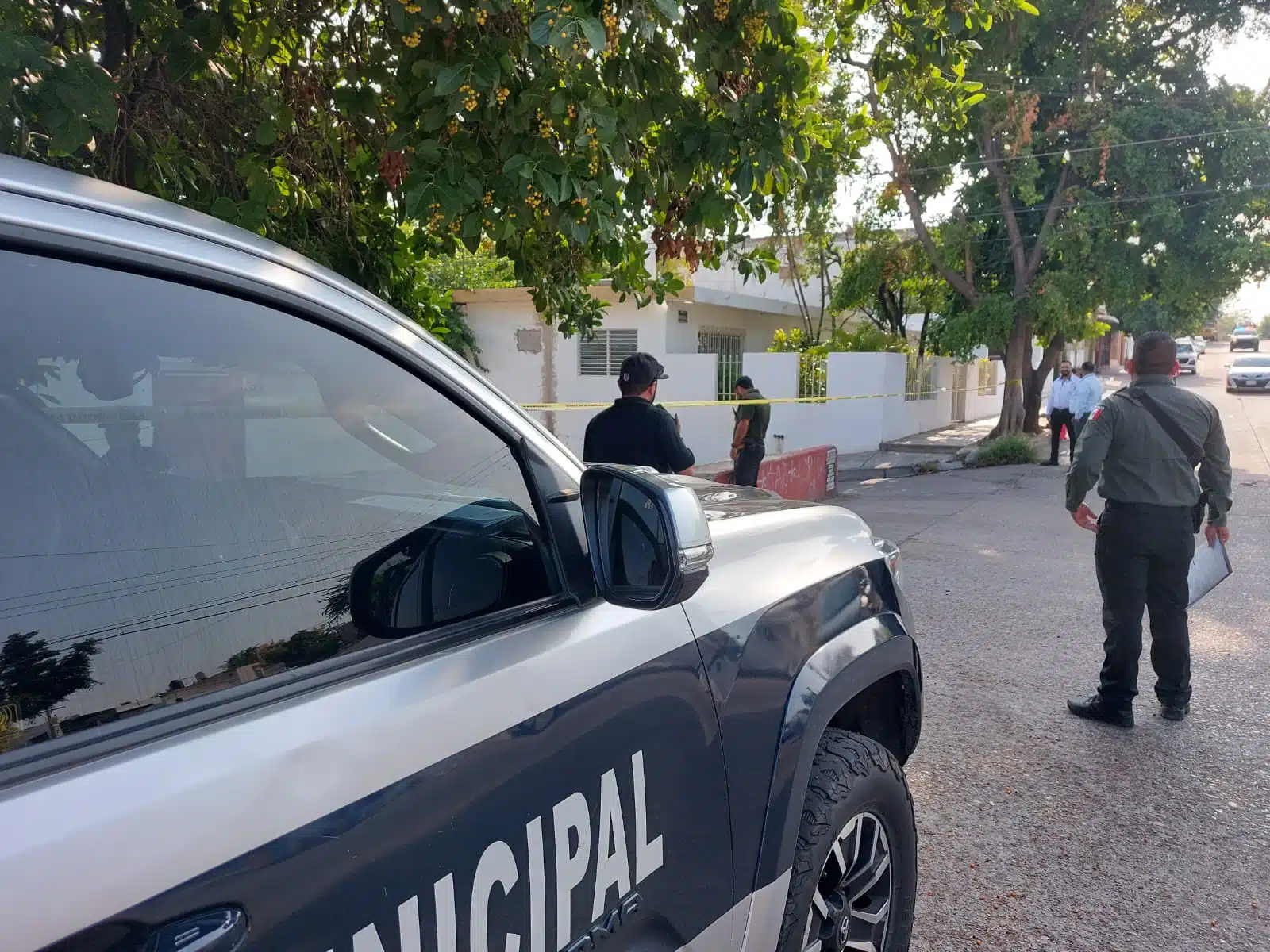 Parte delantera de una camioneta de la policía, 5 personas, árboles, paredes de una casa y carros al fondo