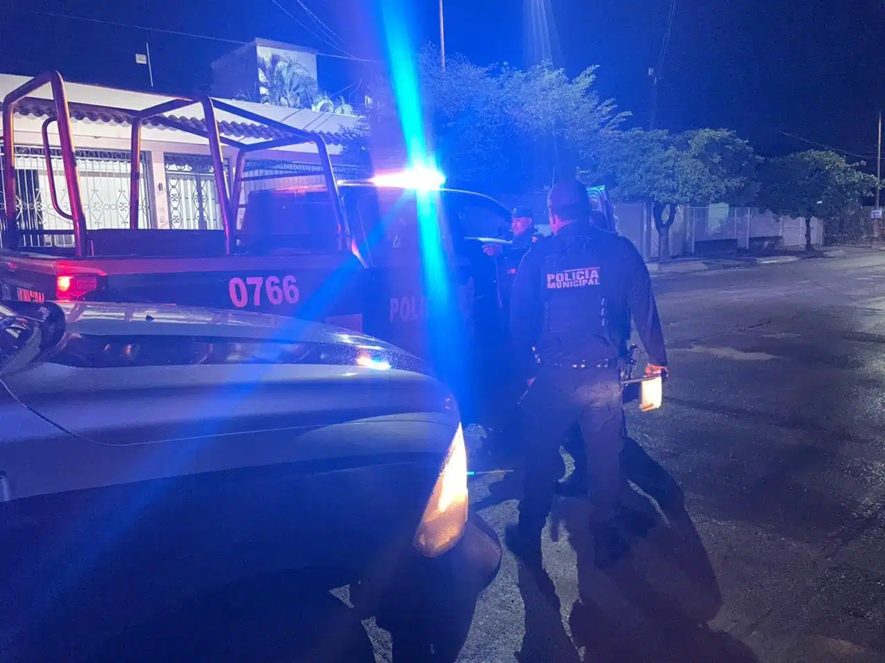 Una camioneta con luces encendidas en la parte de arriba color azul y el frente de otra con la luces prendidas estacionadas sobre una calle, y 2 personas paradas