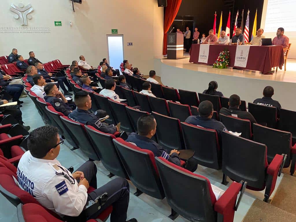 Personas sentadas en un auditorio, una mesa, banderas, paredes y una pantalla proyectora